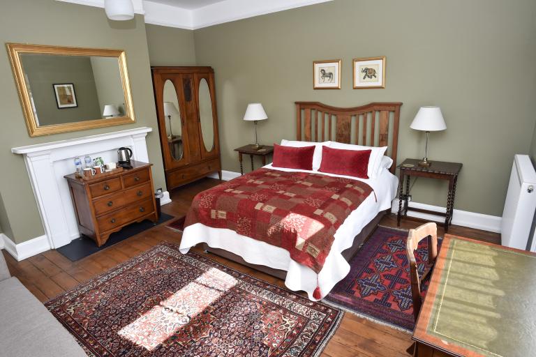Bedroom with wooden furniture, double bed with red patterned bedding, fireplace mantel, rugs on wooden floor, and a wardrobe and bedside tables against neutral walls.