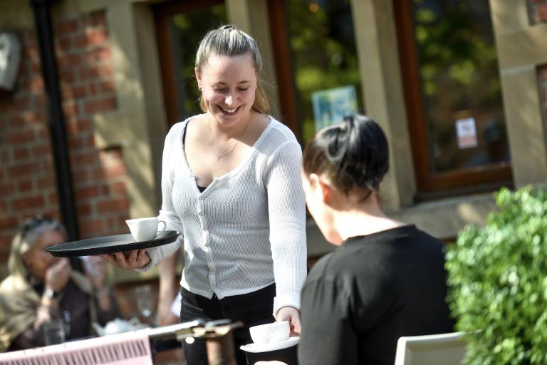 Person serving a hot drink to a seated customer at an outdoor café table.