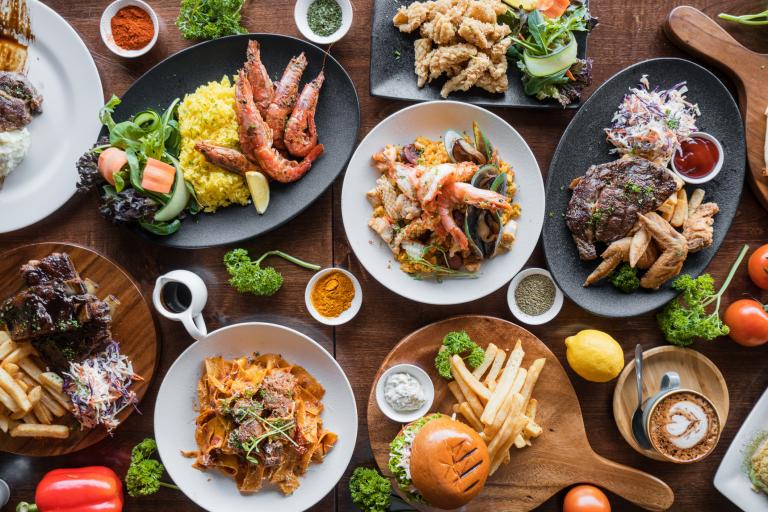 Overhead view of a table filled with dishes including grilled seafood, steak with chips, pasta, burgers and salads, surrounded by small bowls of spices and fresh herbs.