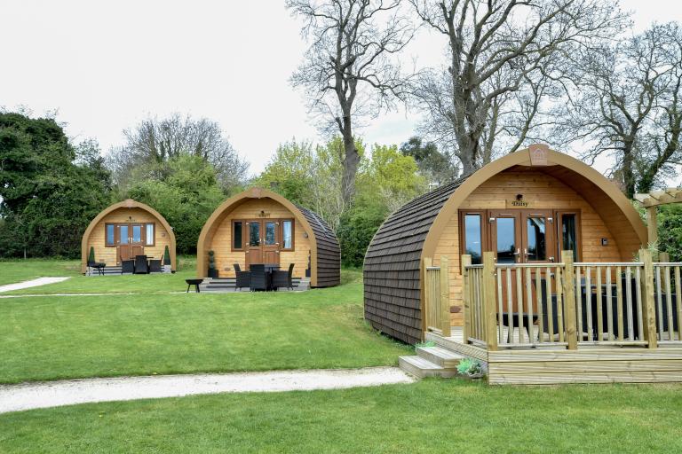 Wooden glamping pods on a grassy site, each with decking and outdoor seating, set among trees along a paved path.