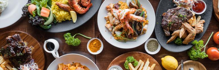 Overhead view of a table filled with dishes including grilled seafood, steak with chips, pasta, burgers and salads, surrounded by small bowls of spices and fresh herbs.