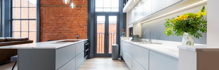 Modern kitchen with exposed brick walls, tall arched windows, and sleek white cabinetry accented by pendant lights and flowers.